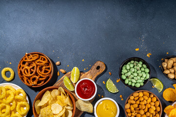 Various unhealthy salty bar snacks set. Dark grey table background with traditional party snacks - chips, onion rings, salted nuts, crisps,  pretzels and sauces top view copy space