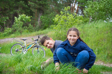 In summer, a brother and sister sit on the grass in the forest.