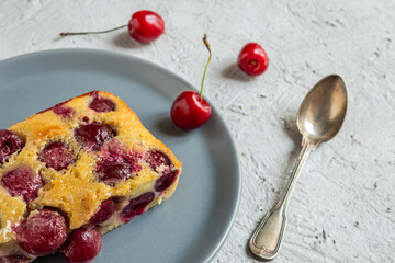 A slice of clafoutis french cherry pie in a gray plate and a spoon on the gray table, top view