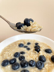 Ripe dark blue blueberries lie in a heap in a white round plate on top of boiled oatmeal, close-up. Plate with food on a beige background. Above the plate is a metal spoon with oatmeal and berries.
