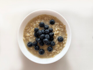 Ripe dark blue blueberries lie in a heap in a white round plate on top of boiled oatmeal, close-up, top view. Plate with food on a beige surface. A shadow falls from the plate to the surface.