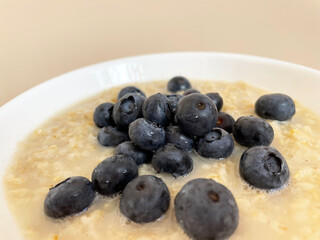 Ripe dark blue blueberries lie in a pile in a white round plate on top of boiled oatmeal, close-up. Plate with food on a beige background. Delicious and healthy breakfast. Vitamins. Proper nutrition.