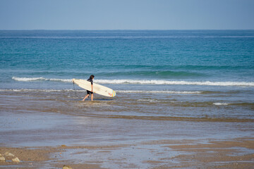 Fototapeta na wymiar Une surfeuse, avec sa planche sous le bras, marche dans l'eau, sur une plage