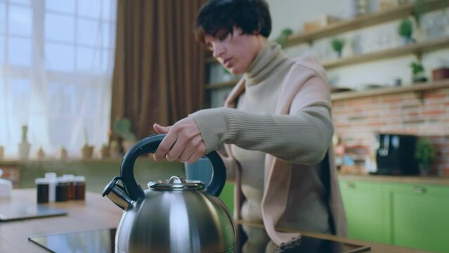 At the kitchen island woman take the kettle from the electric stove and add some boiling water on the cup of tea