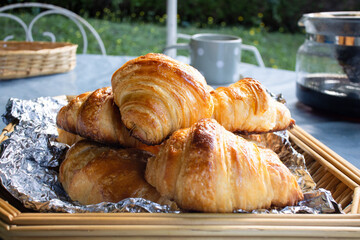 A traditional homemade French butter croissant in a food wooden basket on a table with cup and coffee pot, surrounding with blurry bokeh background of green garden and flowers 