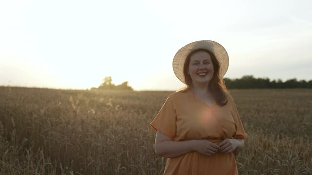 Attractive plus size woman walks through dry wheat field at sunset. Lady in orange dress, hat smiles at camera, enjoys fresh air, unity with nature. Concept of imperfect body, mental health care.