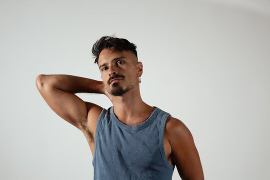 A man lifts his arm up, showing his lean, muscular physique. He is wearing a blue sleeveless shirt. He has a goatee, and earrings.