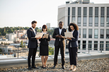 Four investors of caucasian and african american nationality in business outfit standing on rooftop with gadgets and resolving work issues. Each partner looking at own gadget and texting emails.
