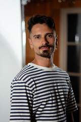 A man poses next to a studio backdrop and looks confidently at the camera. He has a goatee and...