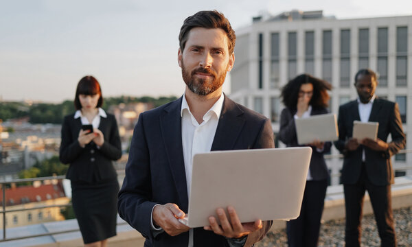 Portrait of cheerful caucasian businessman looking at camera with laptop in hands and smiling. Diverse group of specialists dressed in suits using modern devices for creating project draft drawing.
