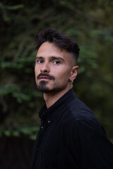 A man looks seriously at the camera, standing in a forest. The background is filled with the greenery of surrounding trees.