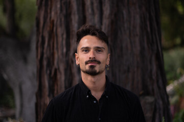 Standing in front of the deeply textured bark of a redwood tree, a man smiles for a portrait. He...