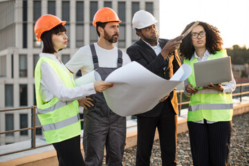 Group of multiethnic industrial employees planning building project and meeting on panoramic terrace. Four international colleagues using computer and technical plan for remodeling business center.