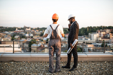 Back view of two male developers wearing hardhats standing on rooftop and talking about apartment...