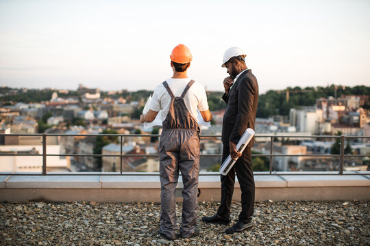 Back view of two male developers wearing hardhats standing on rooftop and talking about apartment facades. Workman in building uniform holding blueprints while business man touching chin and thinking.