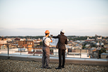 Back view of foreman in grey uniform and business investor in black suit watching together at...