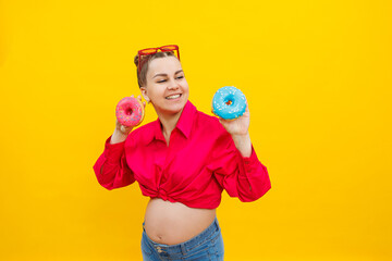 A smiling pregnant woman in a pink shirt holds donuts in her hands on a yellow background. Sweet food during pregnancy. Harmful food during pregnancy.