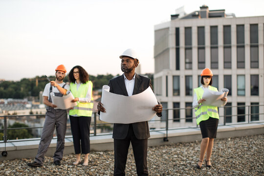 African american businessman wearing suit and hardhat holding project plan in hands and looking aside. Multicultural developers watching at blueprints and discussing construction details.