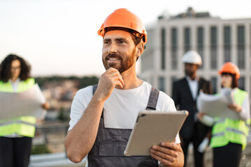 Smiling caucasian builder dressed in uniform and protective helmet holding digital tablet and touching chin with hand. Multicultural group of engineers meeting at terrace on top of construction site.