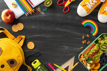 Balanced meal idea for schools. Top view of plastic lunch container featuring natural and healthy food, shoes, backpack, lively stationery on chalkboard backdrop with frame for text or promo content