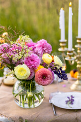 Romantic table decor for a loving couple on a meadow. Two glasses of wine, flowers in a vase, silverware, fruits, chandelier with burning candles, wooden furniture. Sunset, golden hour, summer