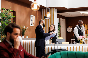 Businessman reviews registration files at hotel reception, talking to front desk staff in lobby upon his arrival. White collar worker entering resort before attending upcoming meeting, business trip.