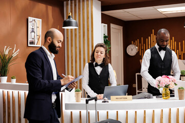 Entrepreneur arrive in hotel reception, signing forms to register and check in. Professional man with formalwear and suitcase staying at luxury resort to attend important business meetings.