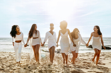 Full length shot of happy ladies walking on the sea shore, holding hands and laughing, having hen...