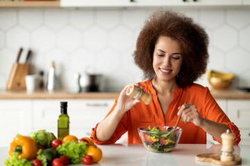 Happy young black lady preparing healthy vegetable salad in kitchen, adding salt