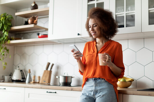 Smiling Black Female Messaging On Smartphone And Drinking Coffee In Kitchen Interior