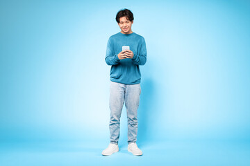 Fototapeta premium Asian guy using smartphone enjoying nice mobile app, blue background