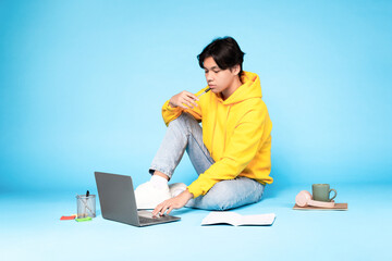 Thoughtful korean teenager guy using laptop sitting on blue background