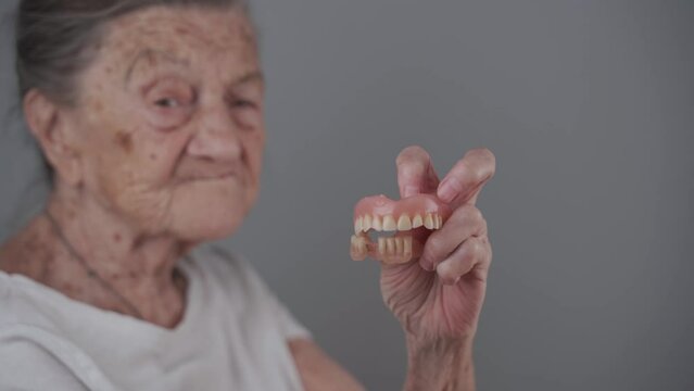 Older woman gets removable dentures, shows on camera. Concept of oral care, false teeth and old age. Full detachable prosthesis in hands of elderly lady with gray hair in studio gray background. 