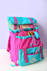 Color school backpack with notebooks, pencil case and stapler on white wooden table near lilac wall