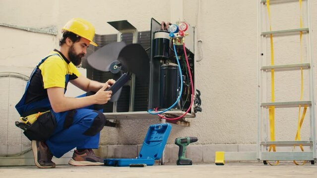 Qualified technician commissioned for outside hvac system routine maintenance, looking for refrigerant issues. Trained engineer doing air conditioner inspection, writing data on tablet