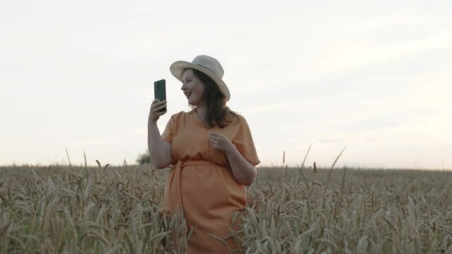 Young attractive woman takes selfie on smartphone in wheat field at sunset. Lady in orange dress and hat smiles, makes duck lips at camera, medium shot. Insects fly near person.