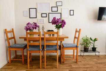 Fototapeta na wymiar Vases with beautiful lilac flowers on table, chairs and blank pictures in interior of light living room