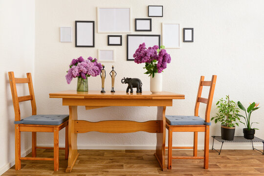 Vases with beautiful lilac flowers on table, chairs and blank pictures in interior of light living room