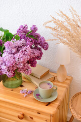 Vase with beautiful lilac flowers, books and cup of coffee on drawers in room, closeup