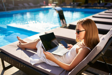 Woman doing remote multitasking work with multiple electronic internet devices on swimming pool beach bed. Freelancer businesswoman telecommuting with tablet, cellphone and laptop from tropical island - obrazy, fototapety, plakaty
