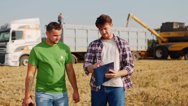 Farmers walk in wheat field, discuss. Agronomist, logistics agent with touch tablet pc negotiate while harvester loading grain to lorry truck. Precision farming with online data management software.