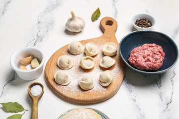 Wooden board with uncooked dumplings and ingredients on white marble background