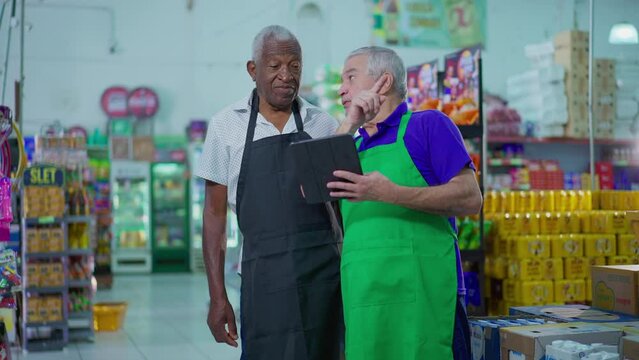 Two senior diverse employees working at supermarket looking at tablet device, teamwork scene of older manager orienting employee at grocery store, job occupation concept