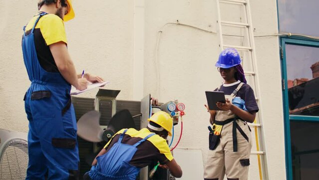 Trained diverse teamworking servicemen cleaning external condenser internal coolants, checking freon level and calibrating thermostats while coworker checks maintenance procedures on tablet