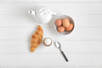 Holder with boiled chicken egg, teapot and croissant on white wooden background