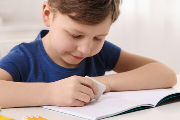 Naklejka premium Little boy erasing mistake in his notebook at white desk indoors