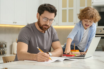 Father working remotely while his son playing with toys at home. Man writing in notebook at desk