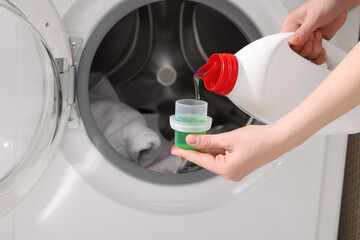 Woman pouring fabric softener from bottle into cap near washing machine, closeup
