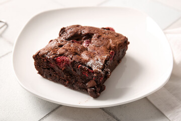 Plate with piece of raspberry chocolate brownie on white tile background