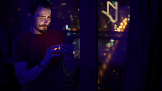 Man is surfing internet on smartphone and knows that he has won the lottery. Young adult caucasian male person standing in dark room next to window using mobile and joying because of good news